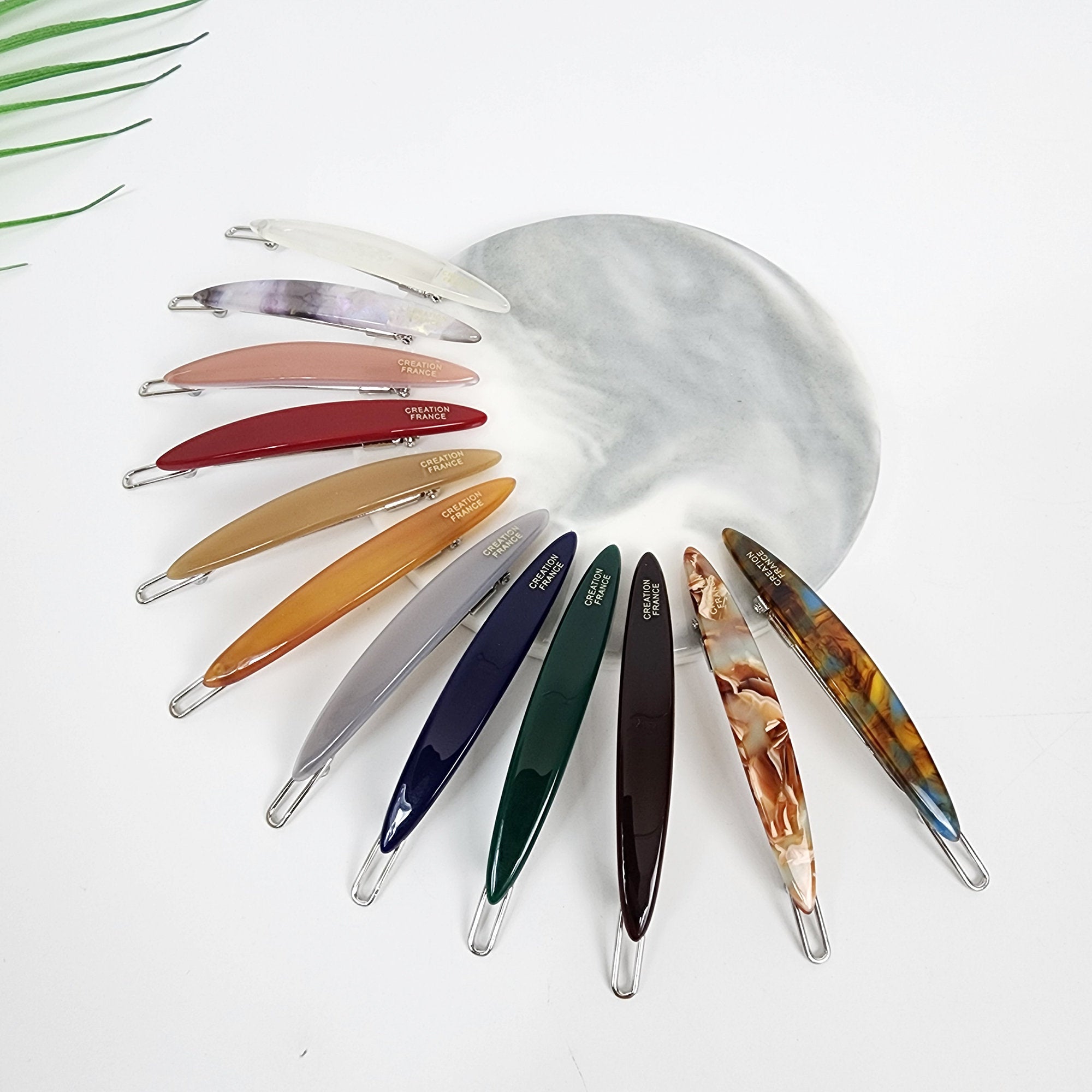 Slim oval shaped cellulose acetate hair clip in a modern minimalist design. Glossy 2.4 inch snap barrette perfect for holding side bangs, available in 12 colors including tortoise and pastel.  A set of twelve hair clips in various colors and patterns, including clear, pink, red, gold, orange, gray, blue, green, brown, and marbled. Each clip has the words 'CREATION FRANCE' printed on it in gold lettering.
