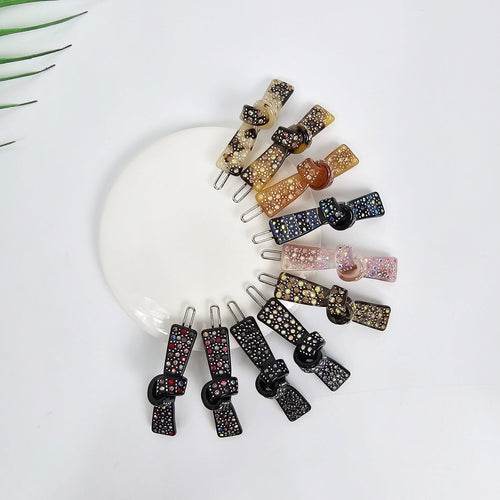 Cellulose acetate flat knot hair clips with rhinestone embellishments. Small 2 inch snap barrettes in tortoise shell and glitter options, perfect for styling bangs or side hair.  A set of ten hair clips with a knot design. The clips are made of black, brown, and white plastic and are decorated with rhinestones. The clips are arranged in a circle on a white surface.