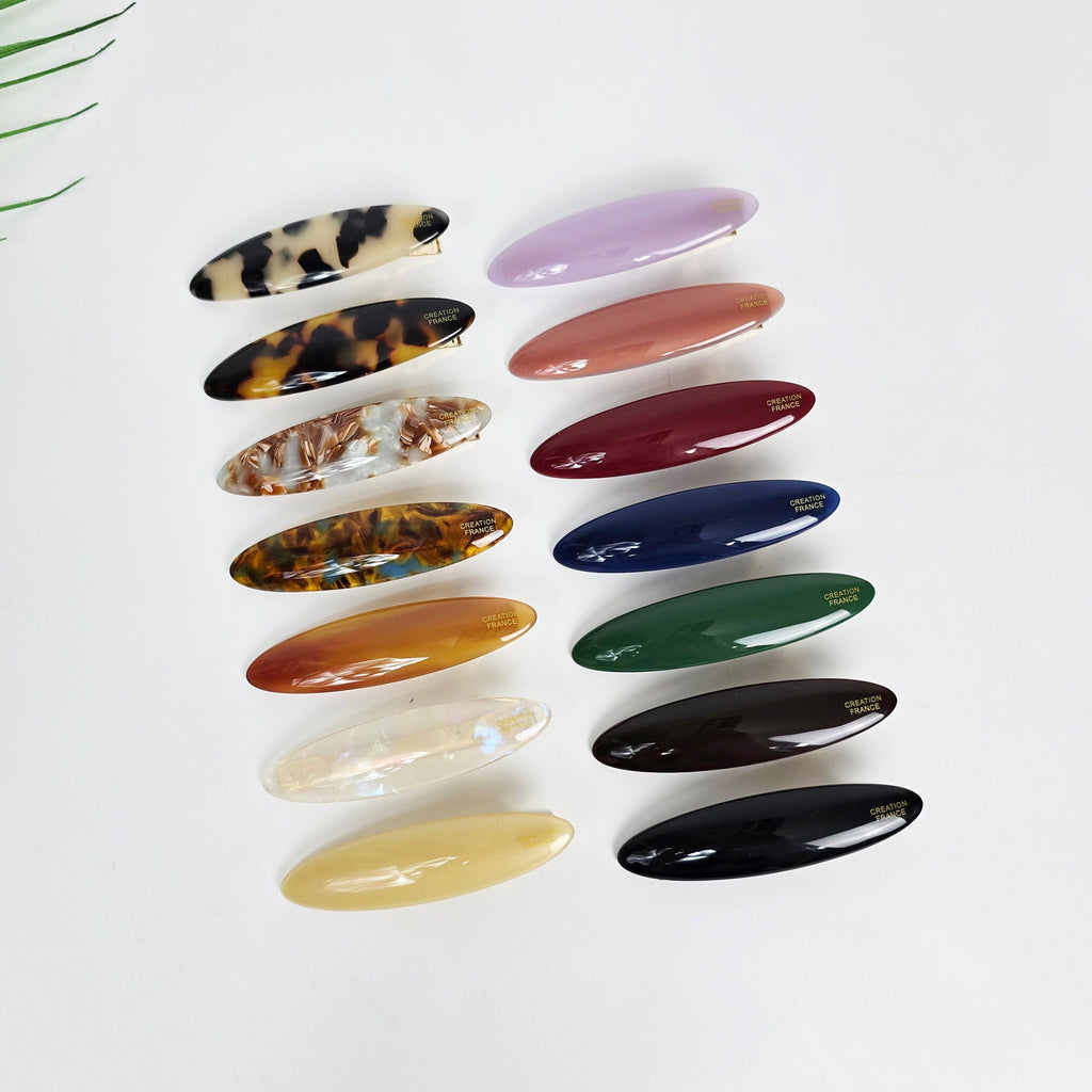 Oval shaped cellulose acetate alligator hair clips in purple, tortoise shell, and solid colors. Sleek 7cm flat beak barrette for women's side bangs styling. Korean minimalist hair accessory. A collection of fourteen assorted color and patterned hair clips. The clips are all oval shaped and have a gold logo that says 'CREATION FRANCE'.