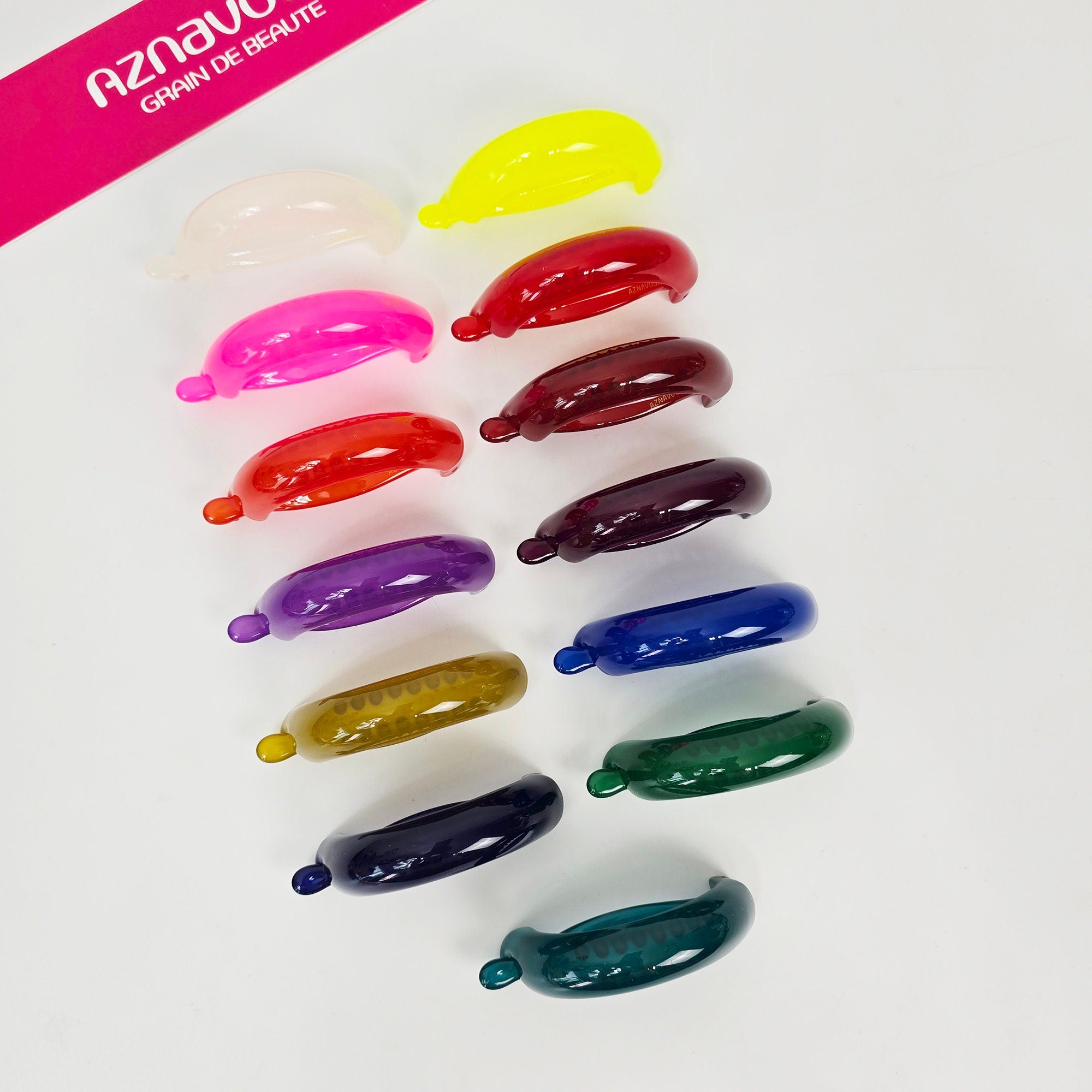 Cellulose acetate curved banana clip in neon pink and glossy colors, small half up hair claw, minimalist ponytail holder for women. Assortment of colorful plastic hair clips in various shapes and sizes. The clips are arranged in a row from yellow to green. The text 'Aznav' and 'Grain de Beaute' is visible in the top left corner of the image.