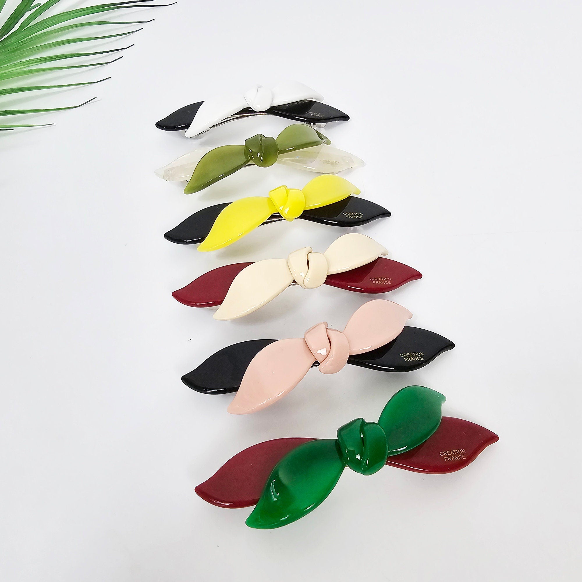 Cellulose acetate hair barrette featuring a two-tone double wave ribbon design in wine and beige. 3.7 inch automatic French clip, perfect for securing side bangs or half-up styles. A set of seven hair clips with bow designs. The clips are in various colors including white, green, yellow, pink, red, and black. Each clip has a small gold label with the text 'CREATION FRANCE'.