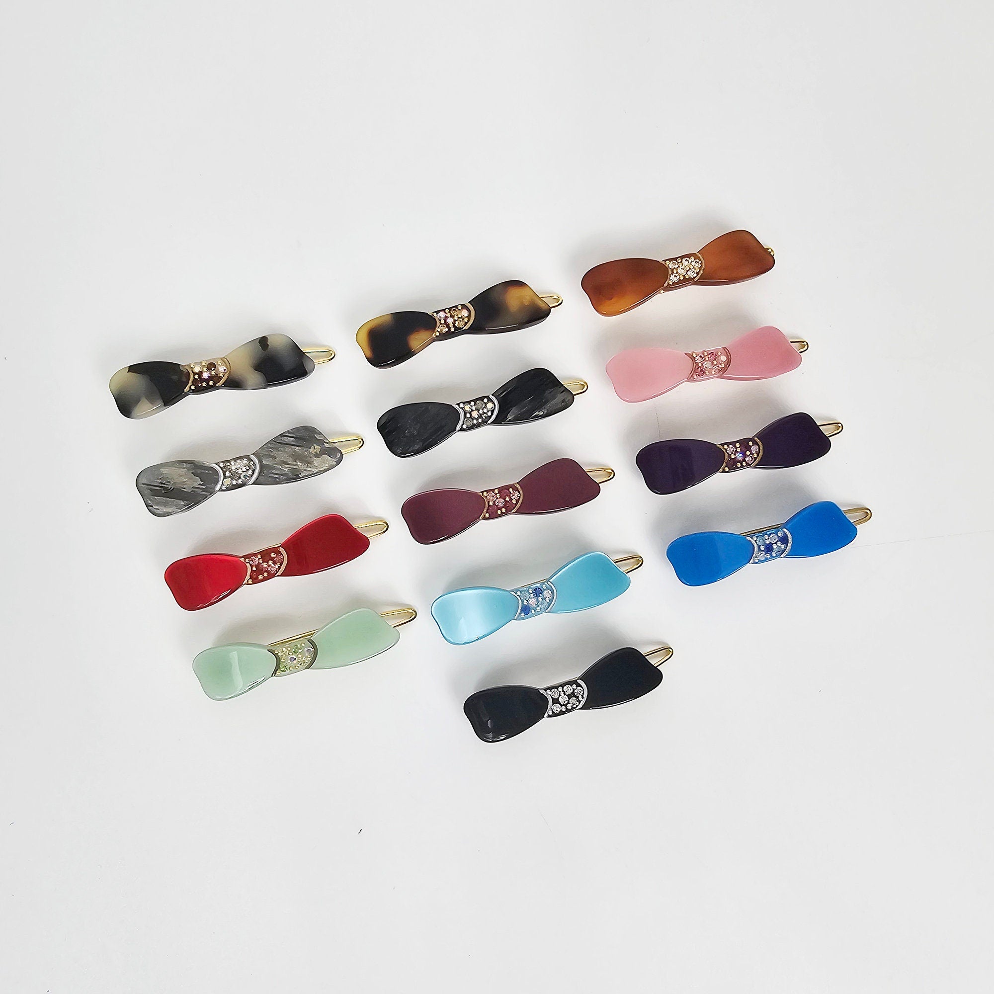 Cellulose acetate small bow hair clip with crystal rhinestone details preppy minimalist style for women and girls bangs. A collection of 15 assorted hair clips with a bow design. The clips are made of plastic and have a variety of colors including black, brown, red, pink, blue, green, and purple. Each clip has a small, sparkly design in the center of the bow.