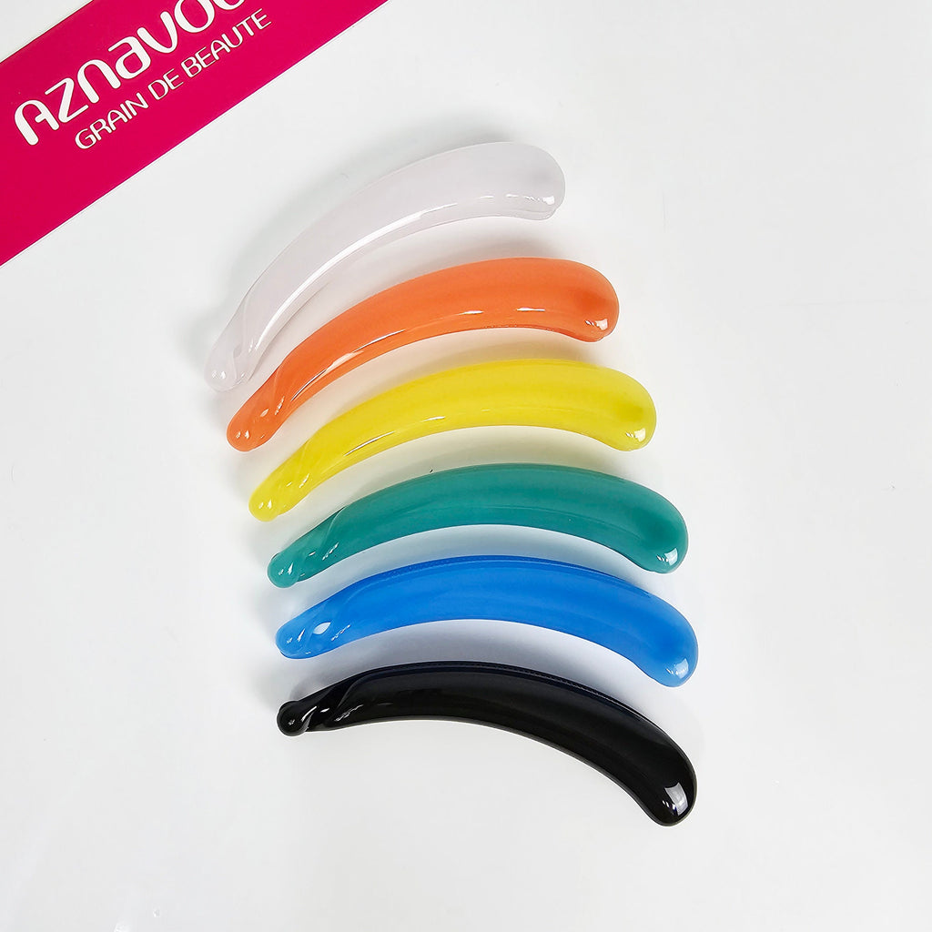Smooth cellulose acetate banana clip in solid colors. 4.3 inch curved vertical hair clamp with inner teeth for a secure ponytail holder on thick hair. Seven curved hair clips in a rainbow of colors: white, orange, yellow, green, blue, black. The clips are arranged in a rainbow arc, with the white clip at the top and the black clip at the bottom.