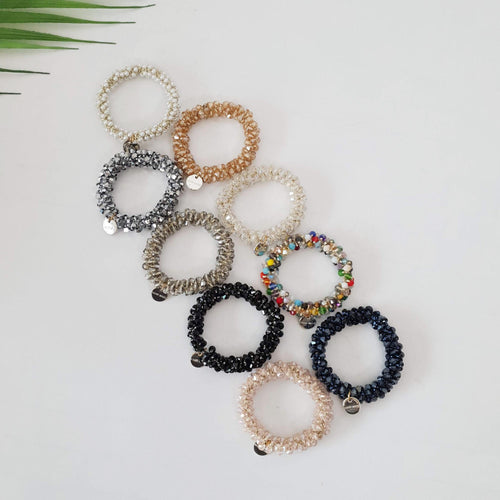 Sparkling glass bead hair tie bracelet with charm, handmade crystal elastic ponytail holder for women. A set of eight beaded hair ties in various colors, including silver, gold, black, and multi-colored. The hair ties are made of small beads and are coiled into a spiral shape. Each hair tie has a small metal tag attached to it.