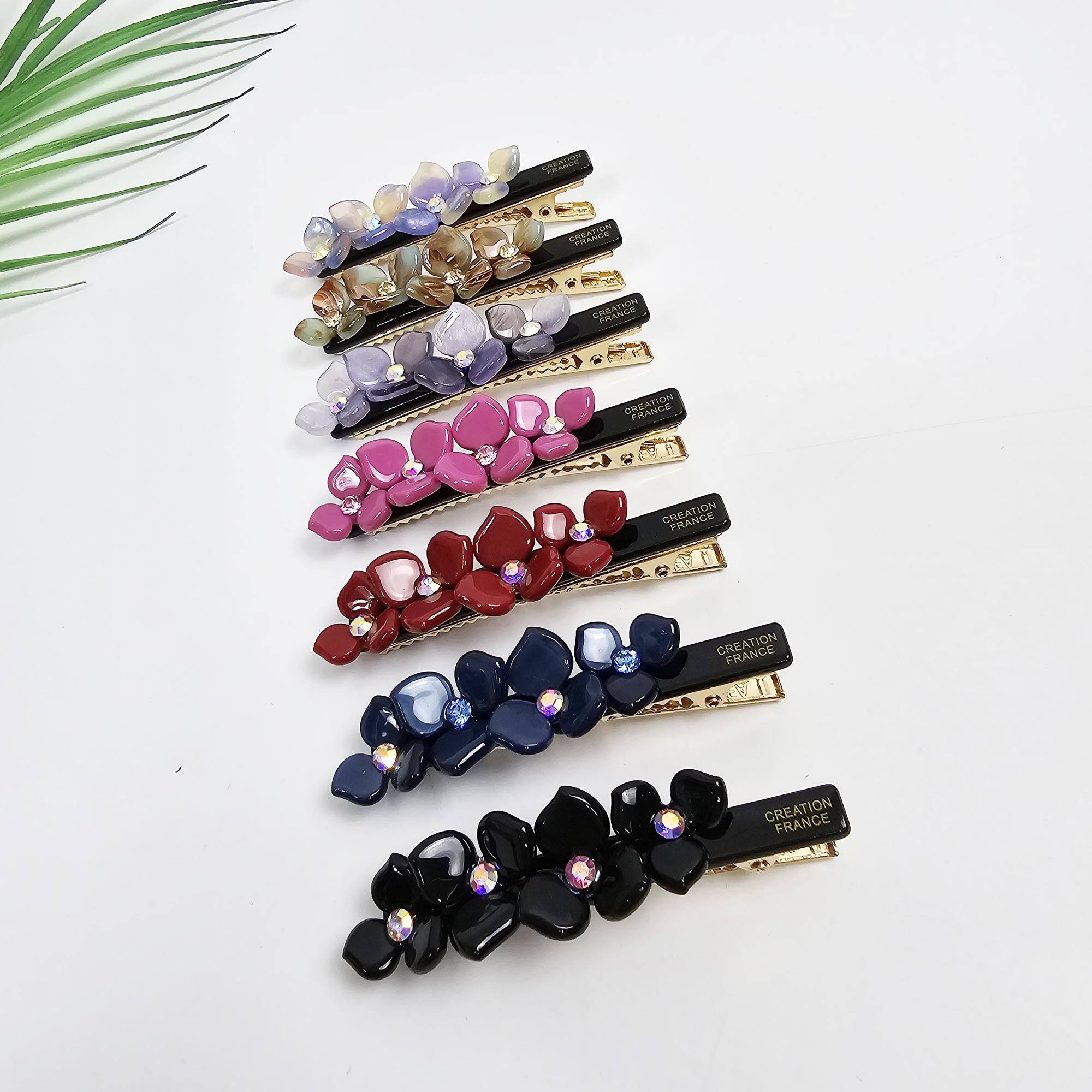 Cellulose acetate floral hair clip with rhinestone centers. 2.7 inch alligator barrette for bangs in pink and tortoise colors. Eight sets of floral hair clips in various colors including lavender, beige, pink, red, navy blue, and black. Each clip features a cluster of resin flowers with sparkling accents, set against a black base.  The gold-toned metal bar clips are marked 'CREATION FRANCE'.