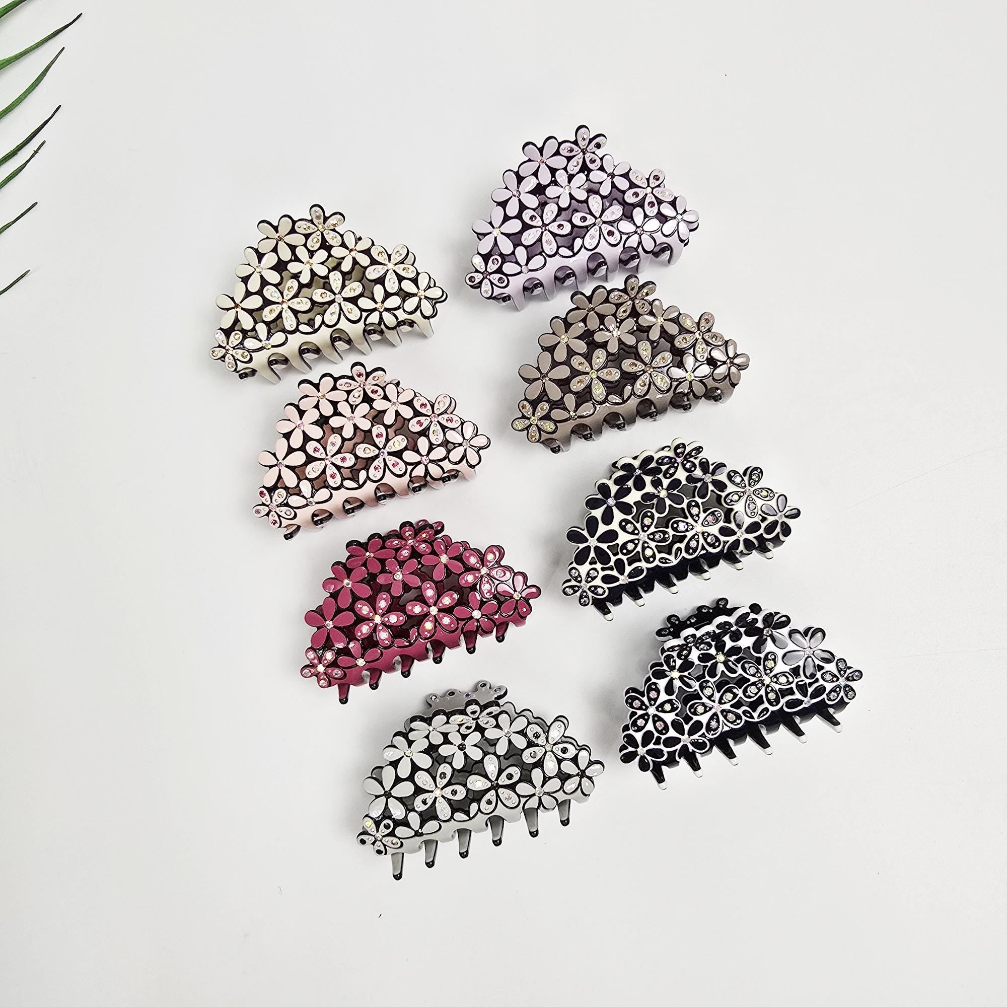 Cellulose acetate flower hair claw clip adorned with rhinestones. 2.6 inch small jaw clip available in pastel colors and black. Perfect for securing half-up hairstyles with a touch of sparkle. Assortment of eight floral hair claw clips in various colors: light purple, beige, pink, burgundy, dark brown, black, and white. Each clip features a half-moon shape adorned with small, flower-shaped embellishments. The clips are displayed against a white background.