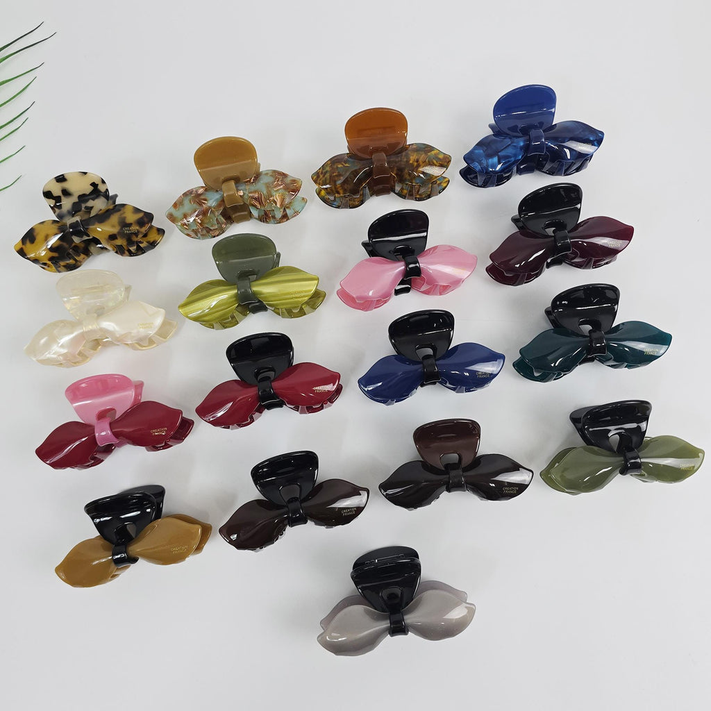 Cellulose acetate ribbon hair claw clip in various colors. 3.3 inch bow design jaw clamp for half-up hairstyles. A collection of 18 plastic hair clips in various colors and patterns. The clips are shaped like a bow and have a textured surface. The clips are arranged in a semi-circle on a white background.