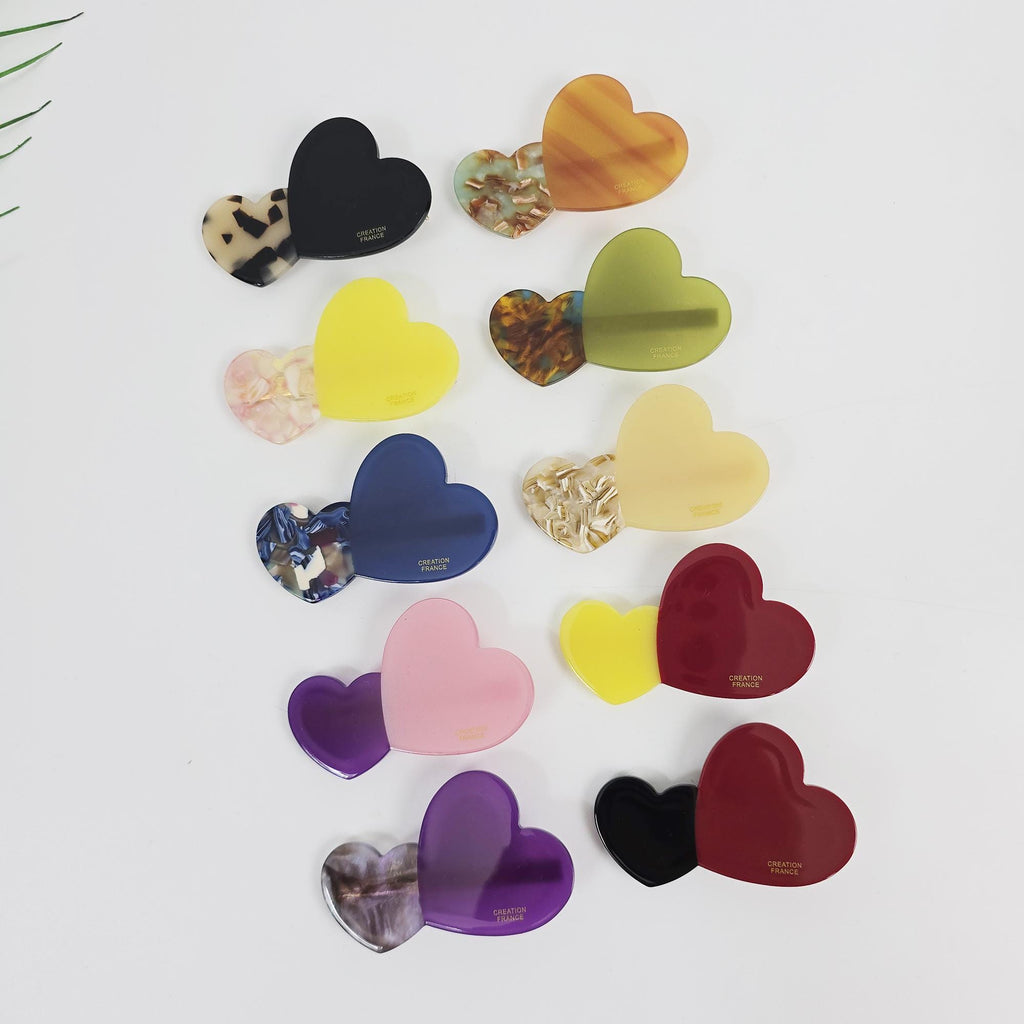 Cellulose acetate double heart hair clip in purple and pink. Small side barrette with gold alligator clip mechanism for girls and women. Twelve heart-shaped hair clips in various colors including yellow, green, red, pink, purple, blue, and black. Each clip has a patterned heart on one side and a solid heart on the other side. The solid hearts have the text 'CREATION FRANCE' printed on them.