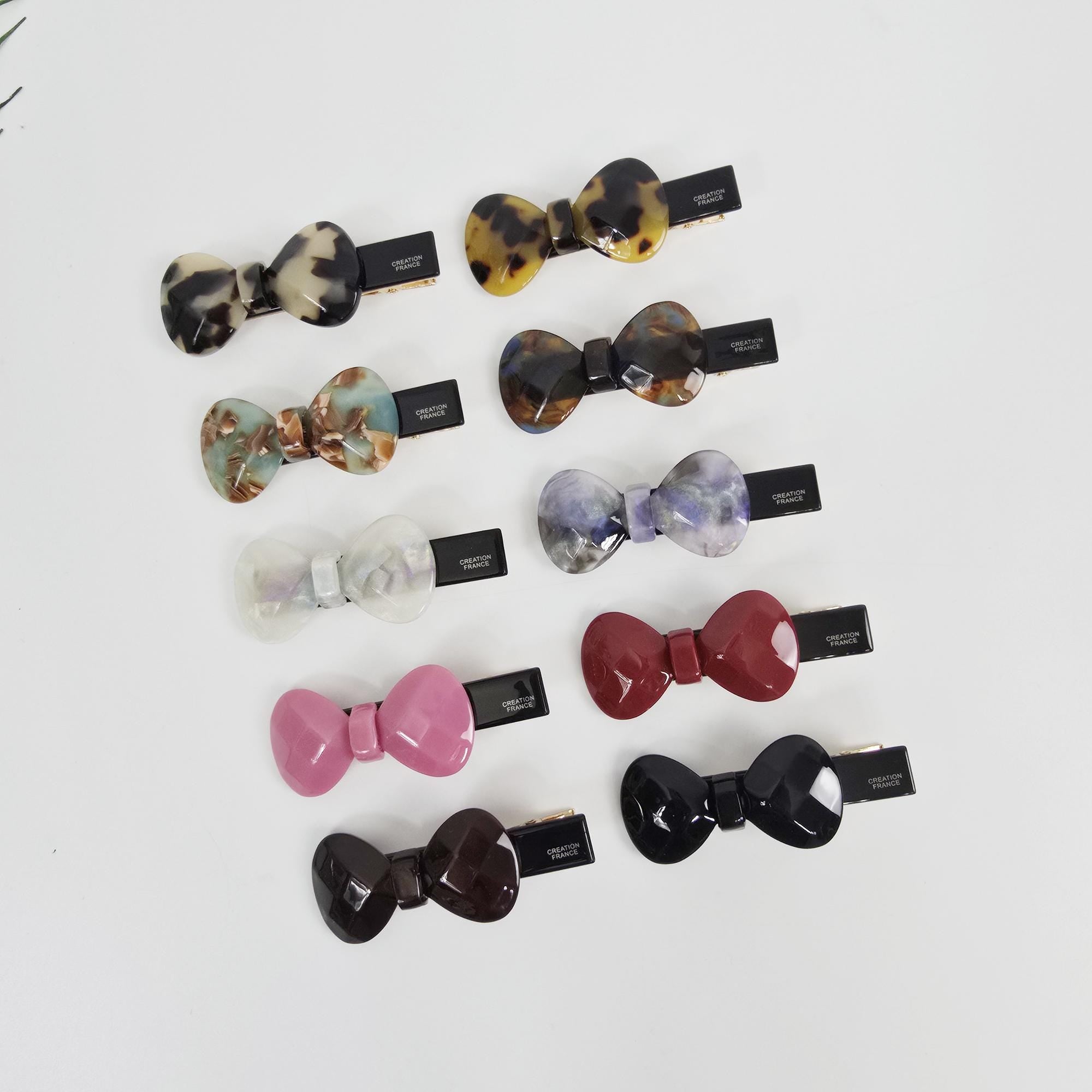 Cellulose acetate bow hair clip featuring tortoise shell and marble patterns. Small side barrette with alligator clip mechanism for girls and women.Ten different colored hair clips in the shape of a bow. The bows are made of a translucent material with different patterns and colors, including black, brown, red, pink, purple, blue, green, and white. The clips are all attached to a black metal clip with the words 'Creation France' printed on them.