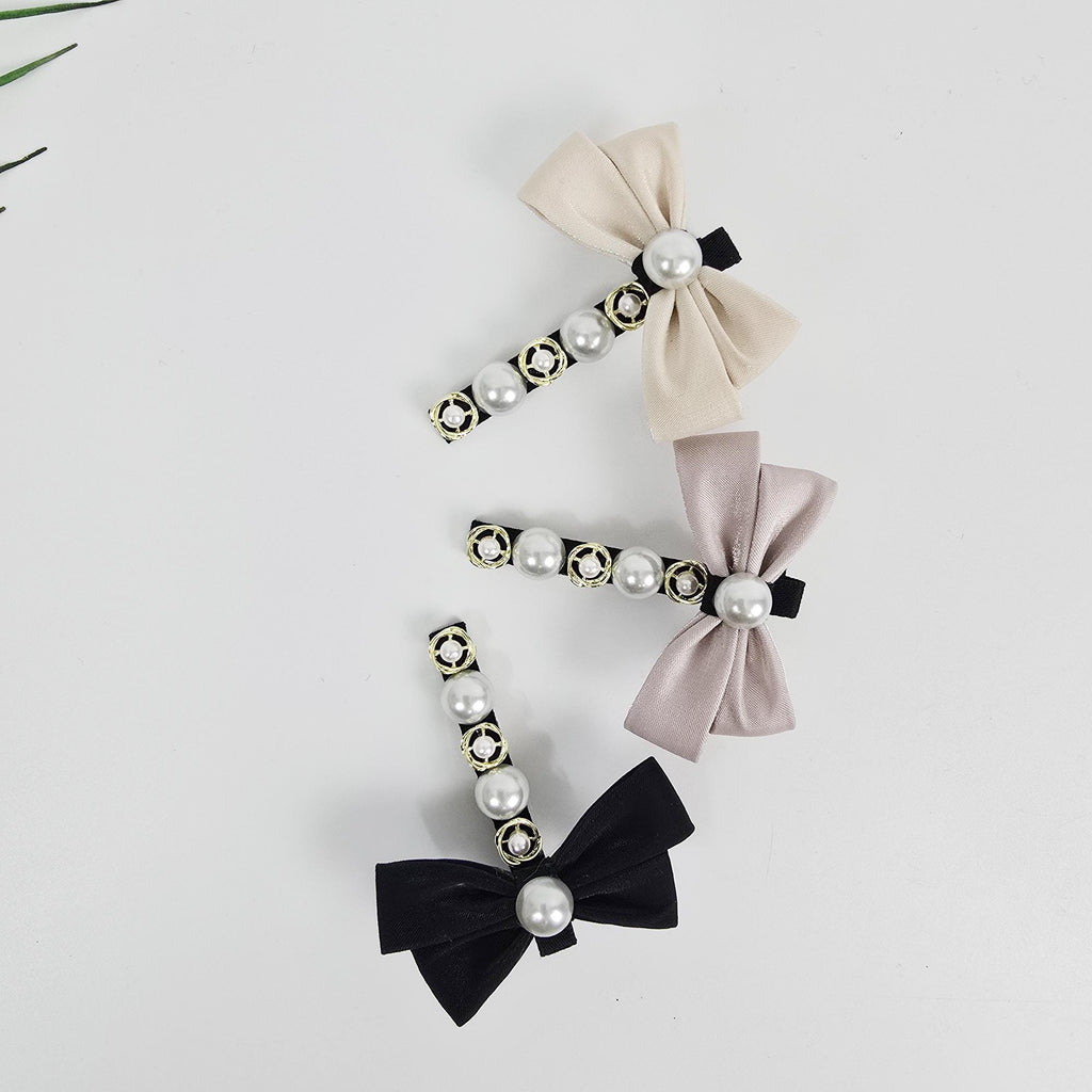 Mini satin bow hair clip with pearl and gold accents. Small alligator side pin for styling bangs or toddler hair in ivory, pink, and black. Three hair clips with pearl and rhinestone embellishments. The clips are black, beige, and light pink. Each clip has a bow made of fabric.
