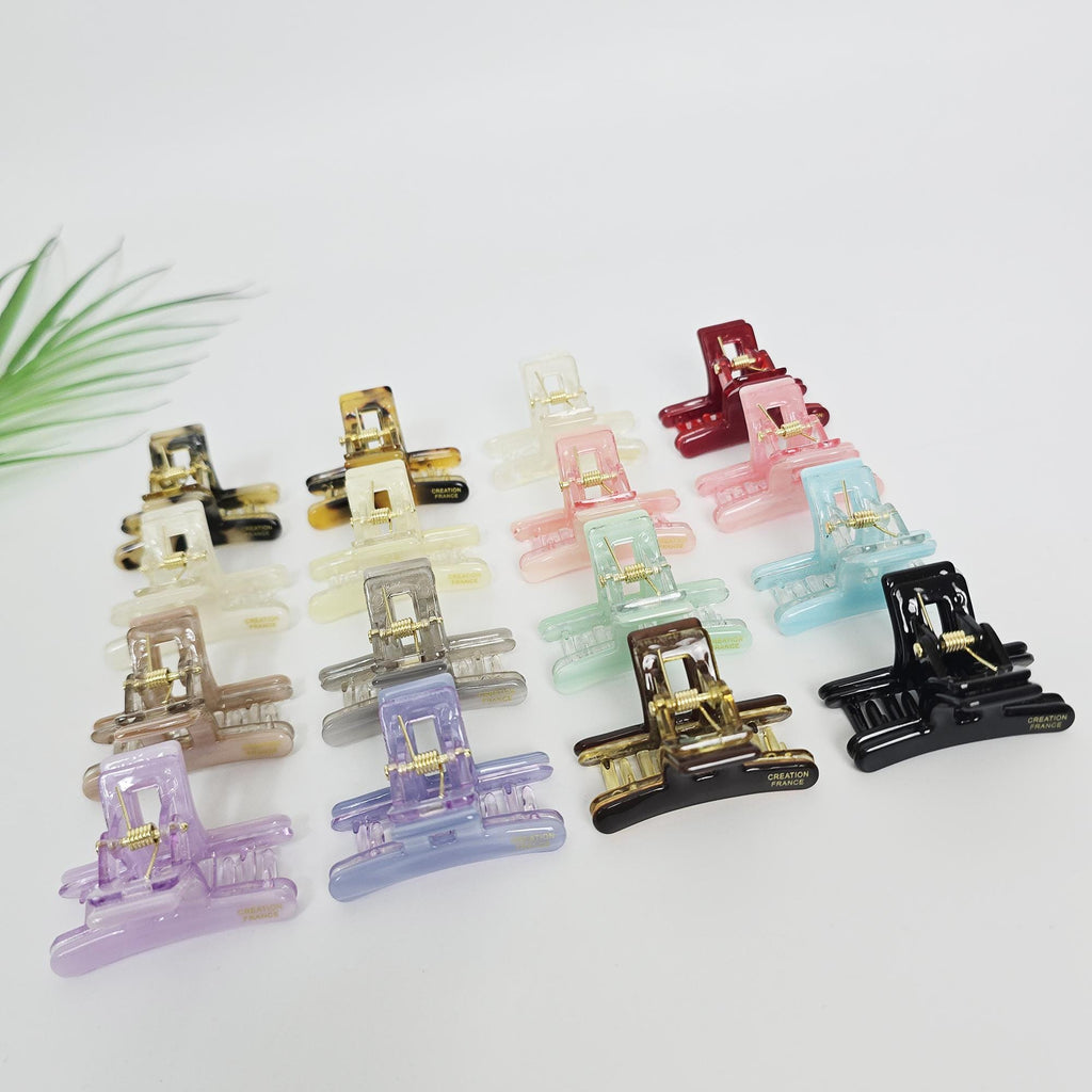 Cellulose acetate rectangular mini hair claw clip with geometric cutout design in pastel and tortoise colors for half-up styling. A collection of small, colorful hair clips. The clips are in various shades including black, brown, clear, gold, pink, red, green, blue, purple, and gray. Each clip has a rectangular shape with a hinge and a decorative element. The clips are arranged on a white surface.