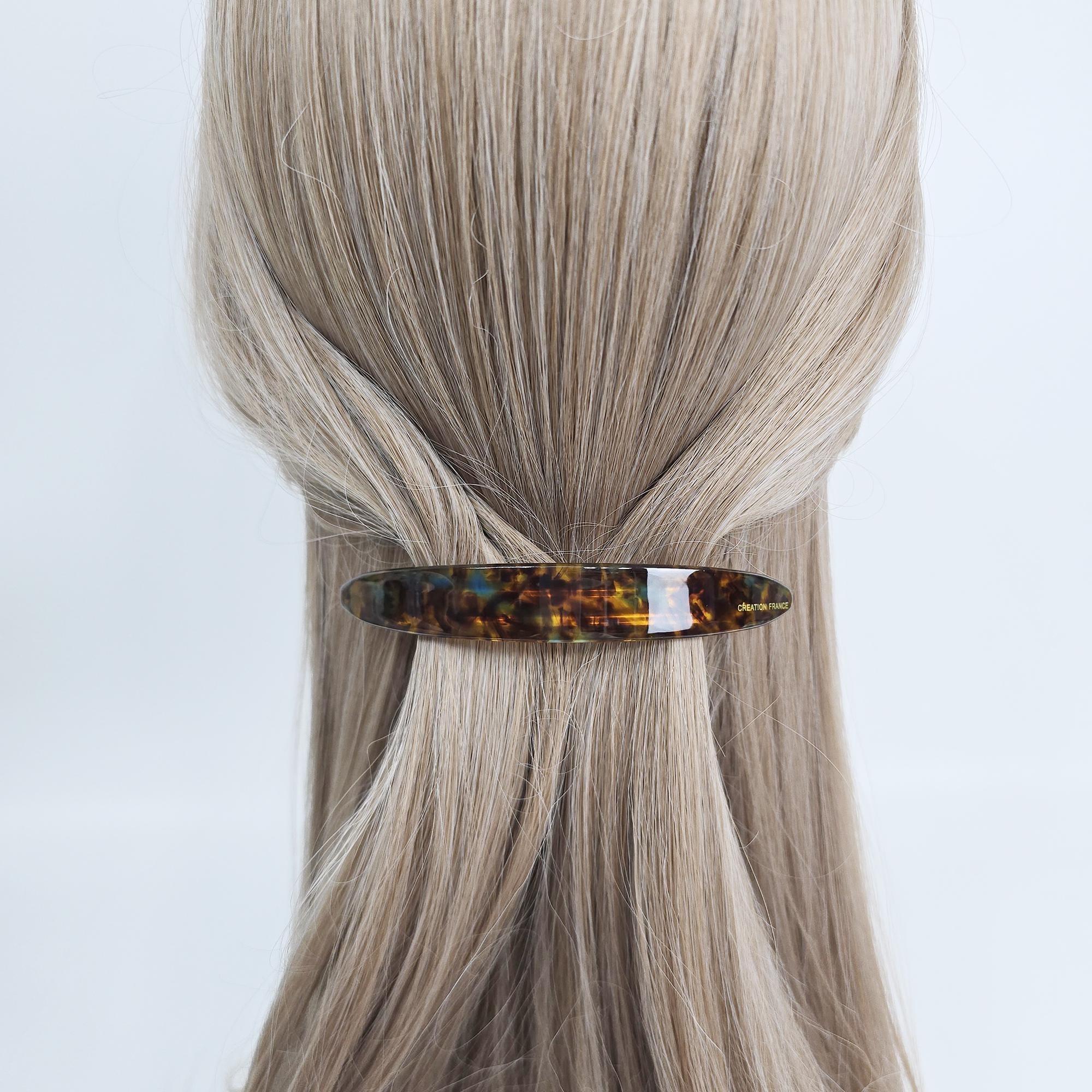 Cellulose acetate long slim curved barrettes in tortoise shell and pastel colors for half up hairstyles. A tortoiseshell hair clip with a rectangular shape. The clip is brown with yellow and black marbling. A white section is in the middle of the clip. The hair accessory is holding back blonde hair.
